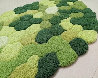 Op de afbeelding: Een groene vloerkleed met een kiezelontwerp. Het vloerkleed heeft verschillende groentinten, van licht tot donker, waardoor een gestructureerd, organisch patroon ontstaat. Het vloerkleed is gemaakt van een zacht, pluche materiaal en is geschikt voor een woonkamer of slaapkamer.