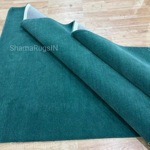 Puede incluir: Una alfombra de color verde azulado, parcialmente enrollada, descansa sobre una superficie de madera de color claro. La textura de la alfombra es visible, y el nombre de la marca "ShamaRugsIN" est&aacute; impreso en la alfombra. La alfombra es de color liso y parece estar hecha de un material suave.