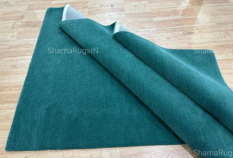 Puede incluir: Una alfombra de color verde azulado, parcialmente enrollada, descansa sobre una superficie de madera de color claro. La textura de la alfombra es visible, y el nombre de la marca "ShamaRugsIN" est&aacute; impreso en la alfombra. La alfombra es de color liso y parece estar hecha de un material suave.