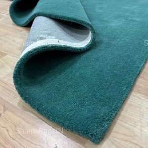 Puede incluir: Una alfombra enrollada de color verde azulado con una textura suave. Se ve la parte inferior de la alfombra, que revela un borde blanco y un respaldo gris. La alfombra se muestra sobre un suelo de madera clara.