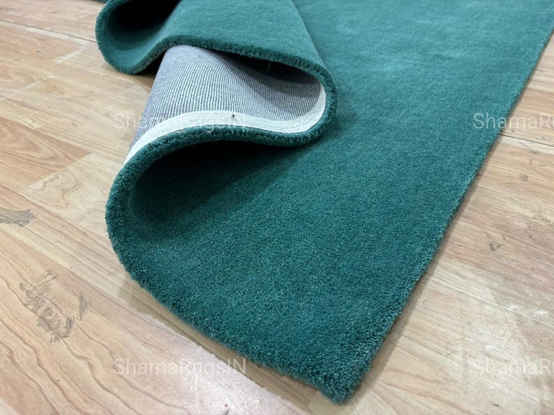 Puede incluir: Una alfombra enrollada de color verde azulado con una textura suave. Se ve la parte inferior de la alfombra, que revela un borde blanco y un respaldo gris. La alfombra se muestra sobre un suelo de madera clara.