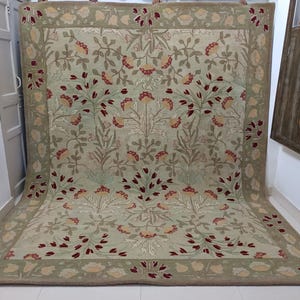 Puede incluir: Una alfombra rectangular grande con un estampado floral en tonos verdes, beige y rojo. La alfombra presenta un borde detallado y un diseño central de flores y hojas estilizadas. La alfombra se exhibe en interiores.