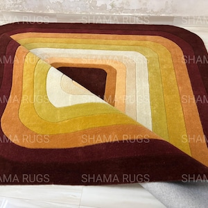 May include: A colorful, geometric rug with a layered design. The rug features a series of concentric rectangles in shades of burgundy, orange, yellow, and cream. The rug is partially rolled up, revealing its underside.