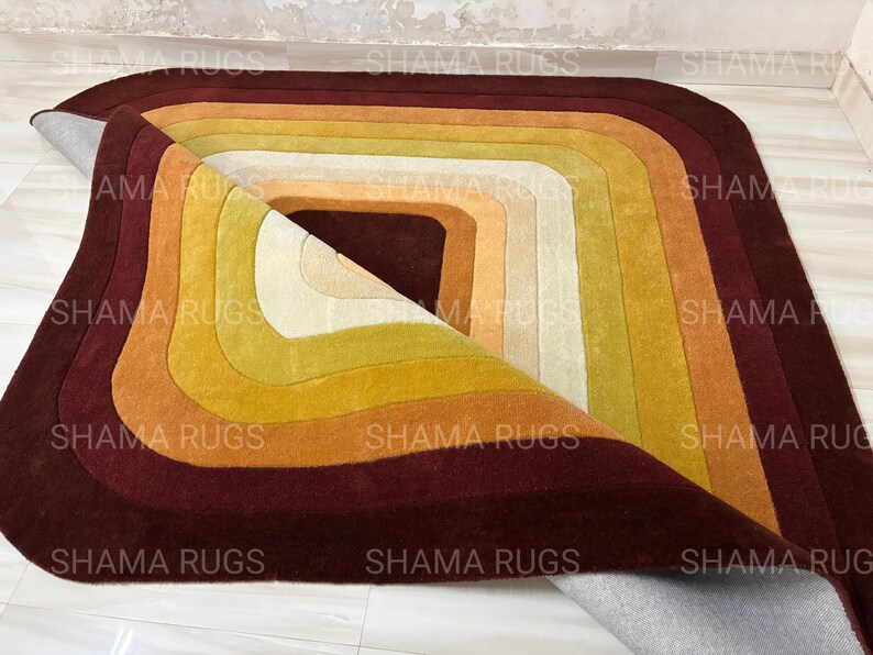 May include: A colorful, geometric rug with a layered design. The rug features a series of concentric rectangles in shades of burgundy, orange, yellow, and cream. The rug is partially rolled up, revealing its underside.