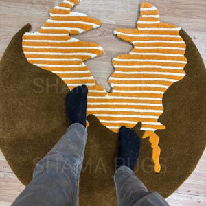 Puede incluir: Una alfombra marrón con un diseño único. La alfombra tiene forma circular con una división en el medio, llena de rayas naranjas y blancas. La alfombra tiene el texto "SHAMA RUGS".
