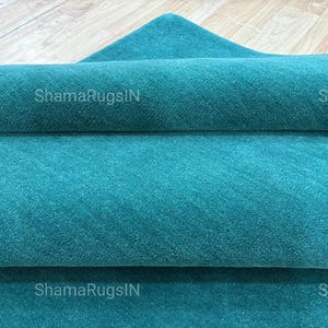 Puede incluir: Una alfombra turquesa enrollada con una superficie texturizada. La alfombra est&aacute; doblada sobre s&iacute; misma, revelando m&uacute;ltiples capas de la tela. El texto "ShamaRugsIN" es visible en la alfombra.