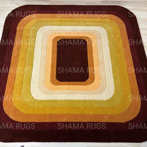 May include: A rectangular rug with rounded corners, featuring concentric layers of color. The rug transitions from a deep burgundy outer edge to a central dark brown square, with shades of gold, yellow, and cream in between. The rug is a decorative floor covering.