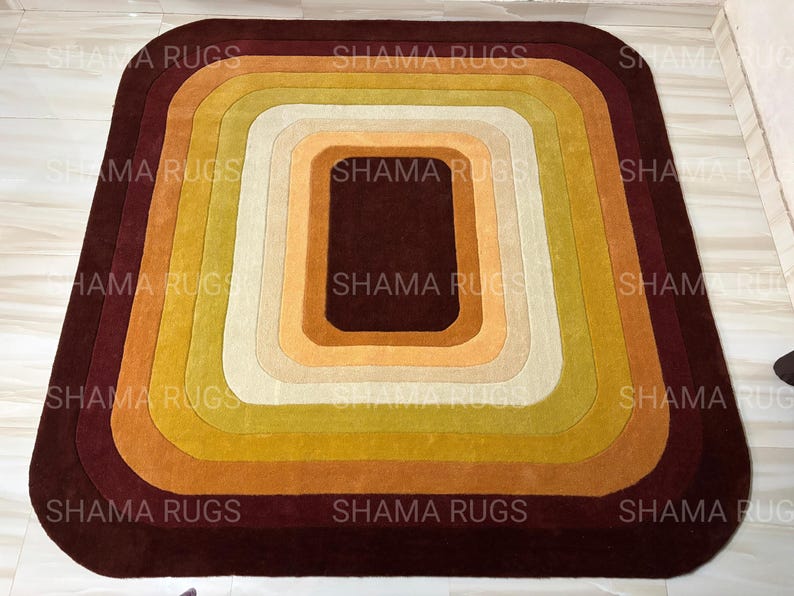 May include: A rectangular rug with rounded corners, featuring concentric layers of color. The rug transitions from a deep burgundy outer edge to a central dark brown square, with shades of gold, yellow, and cream in between. The rug is a decorative floor covering.