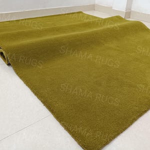 Puede incluir: Una alfombra verde oliva doblada, con una superficie suave y texturizada. La alfombra rectangular parece estar hecha de un material afelpado. Las palabras "SHAMA RUGS" son d&eacute;bilmente visibles en la superficie de la alfombra.