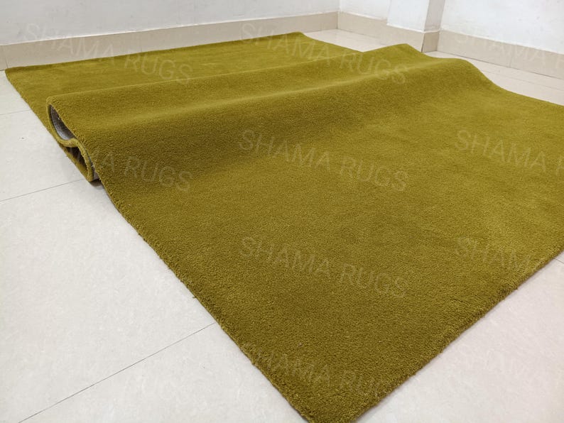 Puede incluir: Una alfombra verde oliva doblada, con una superficie suave y texturizada. La alfombra rectangular parece estar hecha de un material afelpado. Las palabras "SHAMA RUGS" son d&eacute;bilmente visibles en la superficie de la alfombra.