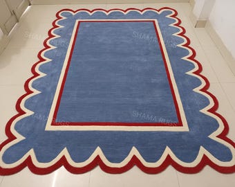 May include: A rectangular rug with a light blue center, framed by a white border and a red scalloped edge. The rug's design is reminiscent of a picture frame, adding a decorative touch to any room. The rug is a floor covering.