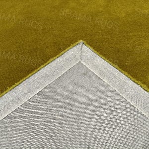 Puede incluir: Primer plano de una secci&oacute;n de una alfombra de color mostaza. Se ve la esquina de la alfombra, que revela un respaldo gris y un borde cosido. La superficie de la alfombra tiene un aspecto suave y texturizado. Las palabras "SHAMA RUGS" son d&eacute;bilmente visibles.