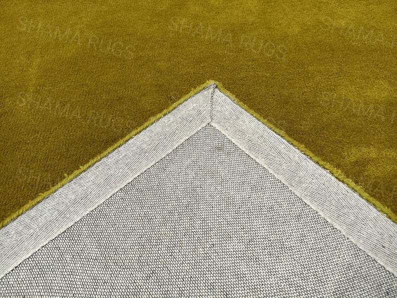 Puede incluir: Primer plano de una secci&oacute;n de una alfombra de color mostaza. Se ve la esquina de la alfombra, que revela un respaldo gris y un borde cosido. La superficie de la alfombra tiene un aspecto suave y texturizado. Las palabras "SHAMA RUGS" son d&eacute;bilmente visibles.