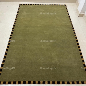 Puede incluir: Una alfombra rectangular con un campo central de color verde oliva sólido. La alfombra está enmarcada por un borde distintivo compuesto por bloques rectangulares alternos en negro y tostado, creando un patrón estructurado en todo el perímetro.