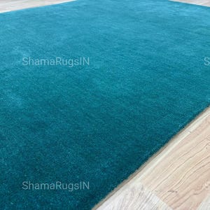 Puede incluir: Una alfombra rectangular de color turquesa con una textura suave y afelpada. La alfombra est&aacute; colocada sobre un suelo de madera clara. El texto "ShamaRugsIN" es visible en la alfombra.