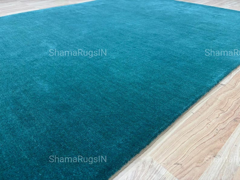 Puede incluir: Una alfombra rectangular de color turquesa con una textura suave y afelpada. La alfombra est&aacute; colocada sobre un suelo de madera clara. El texto "ShamaRugsIN" es visible en la alfombra.