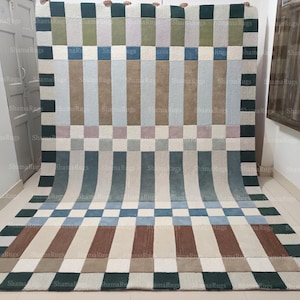 Puede incluir: Una alfombra rectangular con un patrón geométrico. El diseño presenta rayas verticales alternas en tonos de verde, azul, beige y marrón, intercaladas con secciones a cuadros. La alfombra se exhibe sobre un suelo de color claro.