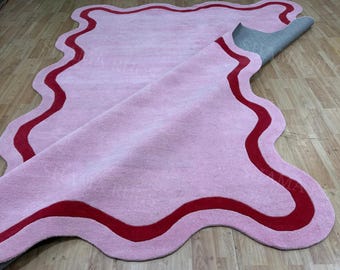 May include: A pink rug with a wavy, irregular shape and a red border. The rug is partially rolled up, revealing a gray backing. The rug is made of a soft, plush material and is suitable for a living room or bedroom.