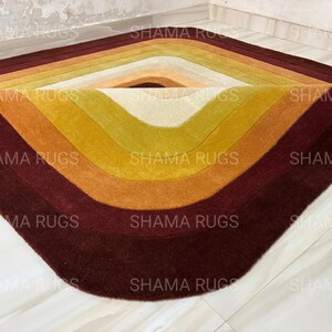 May include: A colorful, geometric rug with a diamond shape. The rug features a gradient of colors, starting with a deep burgundy at the edges and transitioning through shades of orange, yellow, and cream towards the center. The rug is made of a soft material.