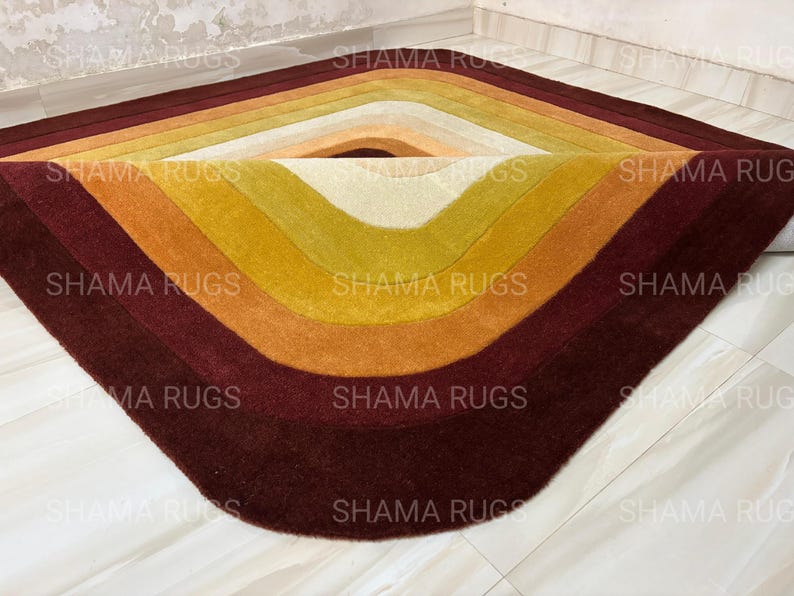 May include: A colorful, geometric rug with a diamond shape. The rug features a gradient of colors, starting with a deep burgundy at the edges and transitioning through shades of orange, yellow, and cream towards the center. The rug is made of a soft material.