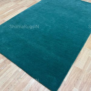 Puede incluir: Una alfombra rectangular de color turquesa con una superficie texturizada. La alfombra se muestra sobre un suelo de madera marr&oacute;n claro. El texto "ShamaRugsIN" es visible en la alfombra.