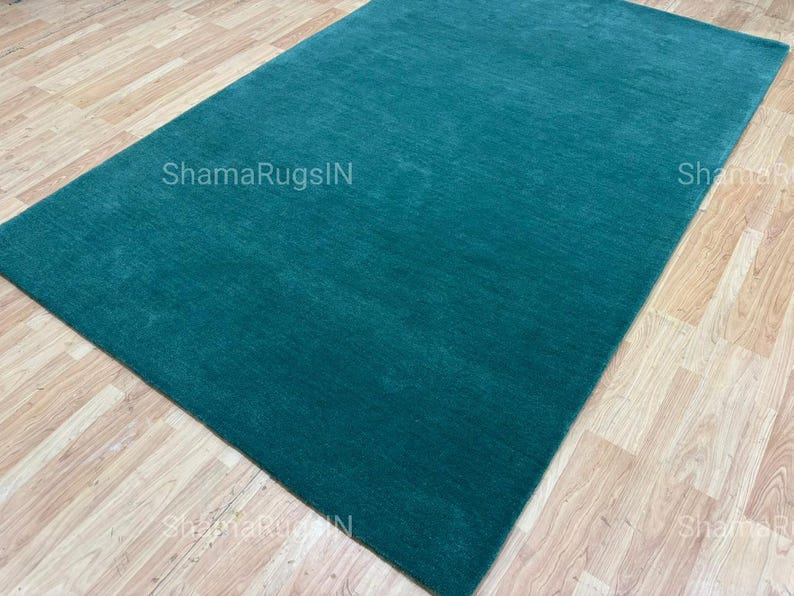 Puede incluir: Una alfombra rectangular de color turquesa con una superficie texturizada. La alfombra se muestra sobre un suelo de madera marr&oacute;n claro. El texto "ShamaRugsIN" es visible en la alfombra.