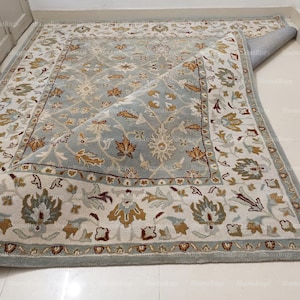 Puede incluir: Una alfombra rectangular con un estampado floral en tonos azul, crema y dorado. La alfombra está parcialmente enrollada, revelando la parte inferior. El diseño presenta un medallón central y un borde detallado.