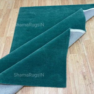 Puede incluir: Una alfombra plegada de color verde azulado con una textura suave y afelpada. La alfombra est&aacute; parcialmente enrollada, revelando un reverso gris. El texto "ShamaRugsIN" es visible en la superficie de la alfombra. La alfombra se exhibe sobre un suelo de madera clara.