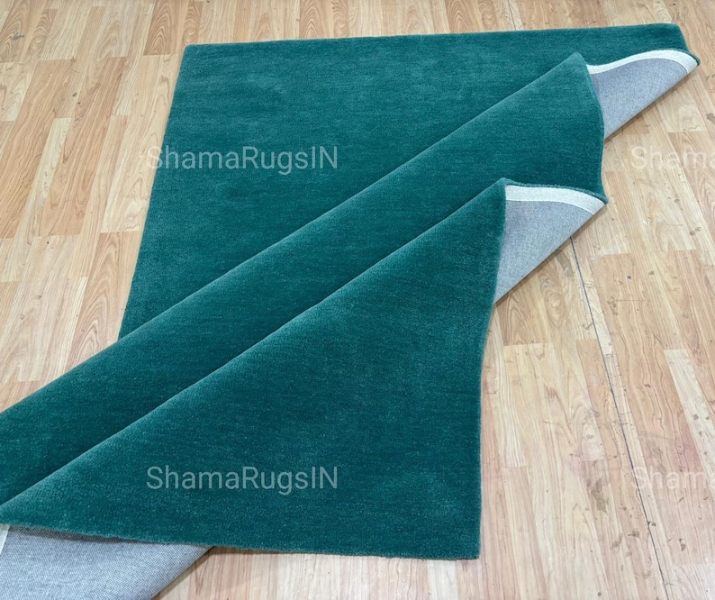 Puede incluir: Una alfombra plegada de color verde azulado con una textura suave y afelpada. La alfombra est&aacute; parcialmente enrollada, revelando un reverso gris. El texto "ShamaRugsIN" es visible en la superficie de la alfombra. La alfombra se exhibe sobre un suelo de madera clara.