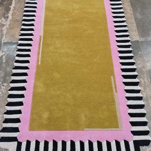 Puede incluir: Una alfombra rectangular con un centro amarillo mostaza, un borde rosa y un borde exterior a rayas blancas y negras. La alfombra tiene un diseño de borde único y festoneado. La alfombra es un revestimiento de suelo decorativo.