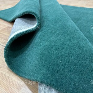 Puede incluir: Una alfombra verde azulado enrollada con una superficie suave y texturizada. Se ve la parte inferior de la alfombra, mostrando un respaldo gris con un borde blanco. La alfombra se exhibe sobre un suelo de madera marr&oacute;n claro.