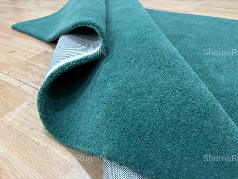 Puede incluir: Una alfombra verde azulado enrollada con una superficie suave y texturizada. Se ve la parte inferior de la alfombra, mostrando un respaldo gris con un borde blanco. La alfombra se exhibe sobre un suelo de madera marr&oacute;n claro.