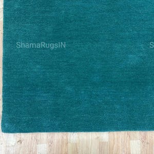 Puede incluir: Una alfombra de color verde azulado con una superficie suave y texturizada. La alfombra es rectangular y parece estar hecha de un material de felpa. Se ve la esquina de la alfombra, con el texto "ShamaRugsIN" impreso en ella. La alfombra est&aacute; colocada sobre un suelo de madera clara.