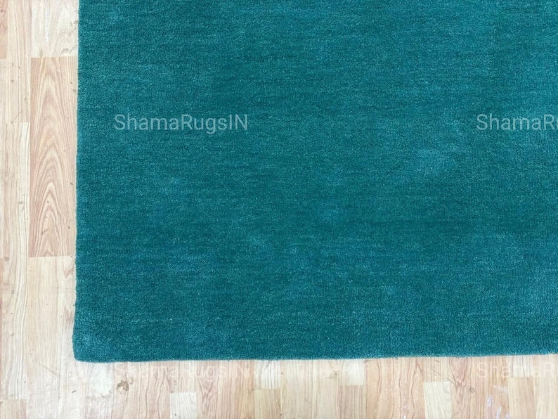 Puede incluir: Una alfombra de color verde azulado con una superficie suave y texturizada. La alfombra es rectangular y parece estar hecha de un material de felpa. Se ve la esquina de la alfombra, con el texto "ShamaRugsIN" impreso en ella. La alfombra est&aacute; colocada sobre un suelo de madera clara.