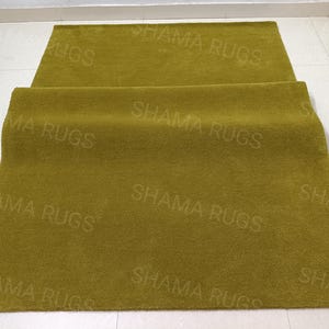 Puede incluir: Dos alfombras verde oliva, una enrollada y la otra extendida. El texto "SHAMA RUGS" est&aacute; impreso repetidamente en la superficie de las alfombras. Las alfombras est&aacute;n sobre un suelo de baldosas blancas.