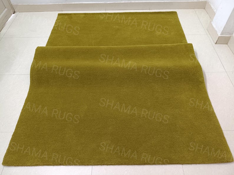 Puede incluir: Dos alfombras verde oliva, una enrollada y la otra extendida. El texto "SHAMA RUGS" est&aacute; impreso repetidamente en la superficie de las alfombras. Las alfombras est&aacute;n sobre un suelo de baldosas blancas.