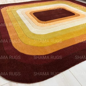 May include: A rectangular rug with a layered design in shades of brown, orange, yellow, and cream. The rug features a central rectangular shape surrounded by concentric curved bands of color. The text "SHAMA RUGS" is repeated across the rug.