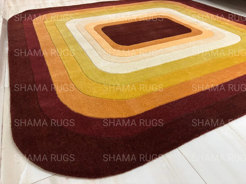 May include: A rectangular rug with a layered design in shades of brown, orange, yellow, and cream. The rug features a central rectangular shape surrounded by concentric curved bands of color. The text "SHAMA RUGS" is repeated across the rug.