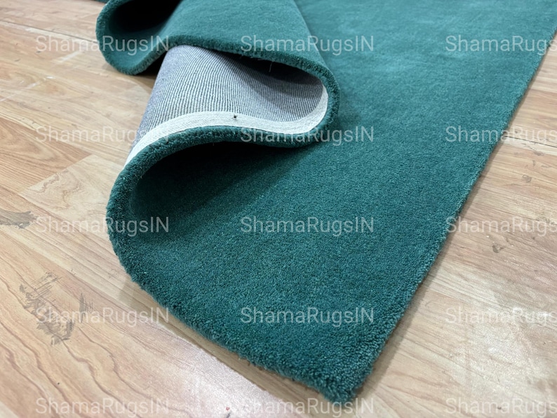 Puede incluir: Una alfombra enrollada de color verde azulado con una textura afelpada. Se ve la parte inferior de la alfombra, mostrando un respaldo blanco con un borde cosido. La alfombra se exhibe sobre un suelo de madera de color claro.
