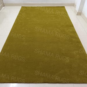 Puede incluir: Una alfombra rectangular de color verde oliva. La alfombra tiene un aspecto suave y texturizado. Las palabras "SHAMA RUGS" est&aacute;n impresas repetidamente en la superficie de la alfombra.