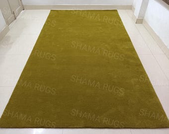 Könnte beinhalten: Ein rechteckiger, olivgrüner Teppich mit dem Text "SHAMA RUGS" wiederholt auf der Oberfläche. Der Teppich liegt auf einem weiß gefliesten Boden, mit einer weißen Wand und einem Türrahmen im Hintergrund.