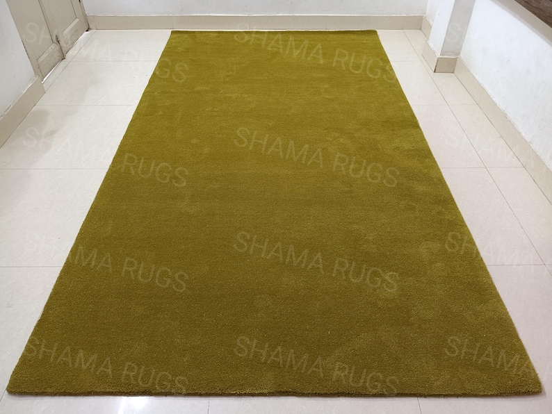 Puede incluir: Una alfombra rectangular de color verde oliva. La alfombra tiene un aspecto suave y texturizado. Las palabras "SHAMA RUGS" est&aacute;n impresas repetidamente en la superficie de la alfombra.