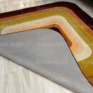 May include: A multi-colored rug with a layered, curved design. The rug features shades of brown, gold, orange, and cream. The underside of the rug is gray. The text "SHAMA RUGS" is repeated across the image.