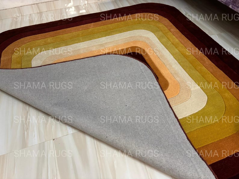 May include: A multi-colored rug with a layered, curved design. The rug features shades of brown, gold, orange, and cream. The underside of the rug is gray. The text "SHAMA RUGS" is repeated across the image.