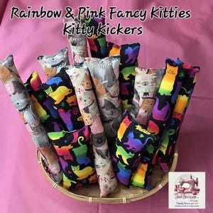 Könnte beinhalten: Eine Sammlung von Katzenspielzeugen, oder "Kitty Kickers", in einem geflochtenen Korb. Die zylindrischen Spielzeuge sind mit bunten Stoffen mit Katzen- und Regenbogenmustern bedeckt. Der Text "Rainbow & Pink Fancy Kitties Kitty Kickers" ist oben zu sehen.