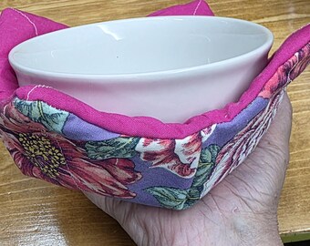 May include: A fabric bowl cozy featuring a vibrant floral pattern with pink, purple, and green tones, constructed with a solid pink fabric border. The cozy holds a plain white ceramic bowl, providing a protective layer for holding hot food items.
