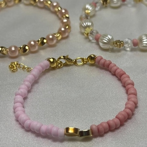 Puede incluir: Primer plano de tres pulseras de cuentas. Las pulseras presentan cuentas rosas y doradas. Una pulsera tiene pequeñas cuentas rosas, otra tiene cuentas rosas más grandes con detalles dorados, y la tercera tiene cuentas transparentes y rosas con detalles dorados.