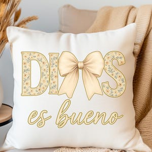 May include: Cream-colored decorative pillow with the words "DIOS es bueno" in gold script. The word "DIOS" is in a floral pattern with a cream-colored bow. The pillow is on a beige couch with a beige blanket.
