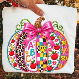 May include: A white t-shirt featuring a large, colorful pumpkin graphic. The pumpkin has segments with leopard print, a checkered pattern, and lined paper with the letters ABC. It is decorated with red apples, yellow pencils, smiley faces, and a large pink bow.