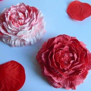 May include: A pair of handmade paper flowers, one in pink and white, the other in red, are the main subjects. Red petals are scattered around them on a light blue surface. The flowers display intricate, layered petal details.